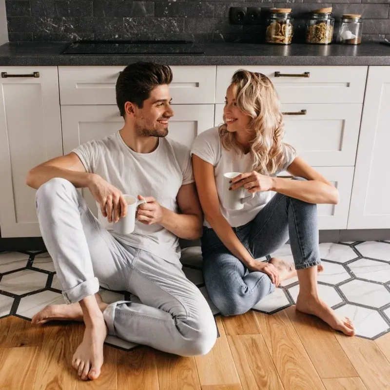 Ein Mann und eine Frau sitzen in einer Küche auf dem Boden und halten Tassen Kaffee in den Händen.