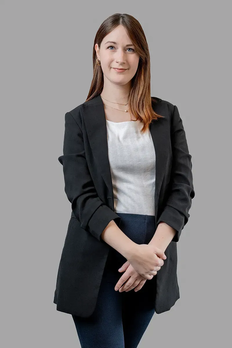 Eine Frau in einer schwarzen Jacke und einem weißen Hemd steht mit gefalteten Händen da.