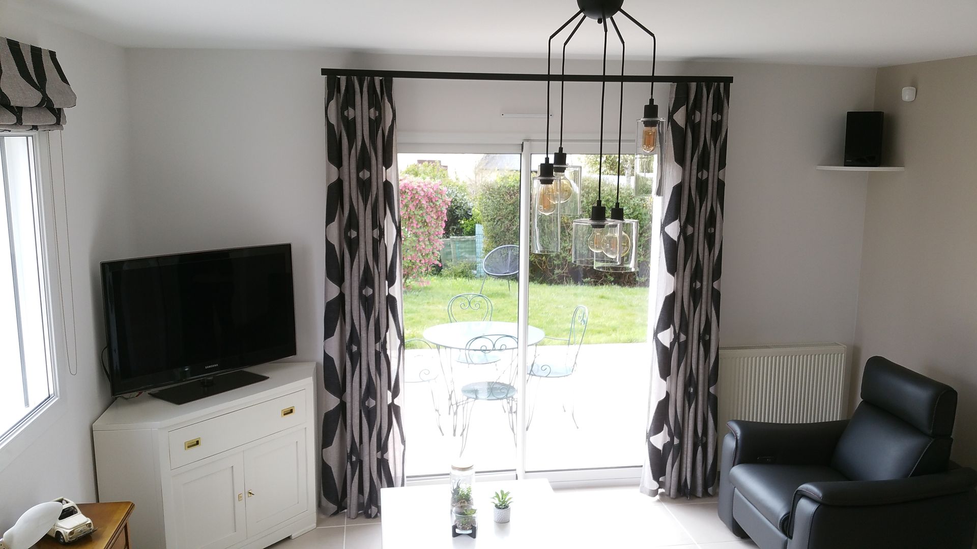 Salon avec meuble blanc de télévision, table basse de couleur blanche, fauteuil cuir noir, devant nous rideaux noirs et gris avec un élégant rail à rideaux noir. Vue sur une terrasse extérieure. A gauche store bateau dans le même tissu que les rideaux