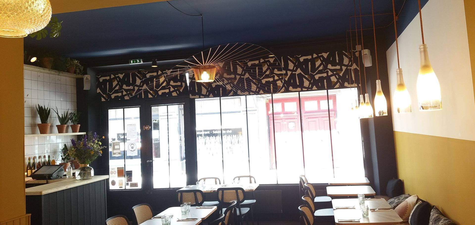 Salle de restaurant avec plafond peint en bleu, murs beige et ocre et stores à motifs géométriques assortis des même couleurs