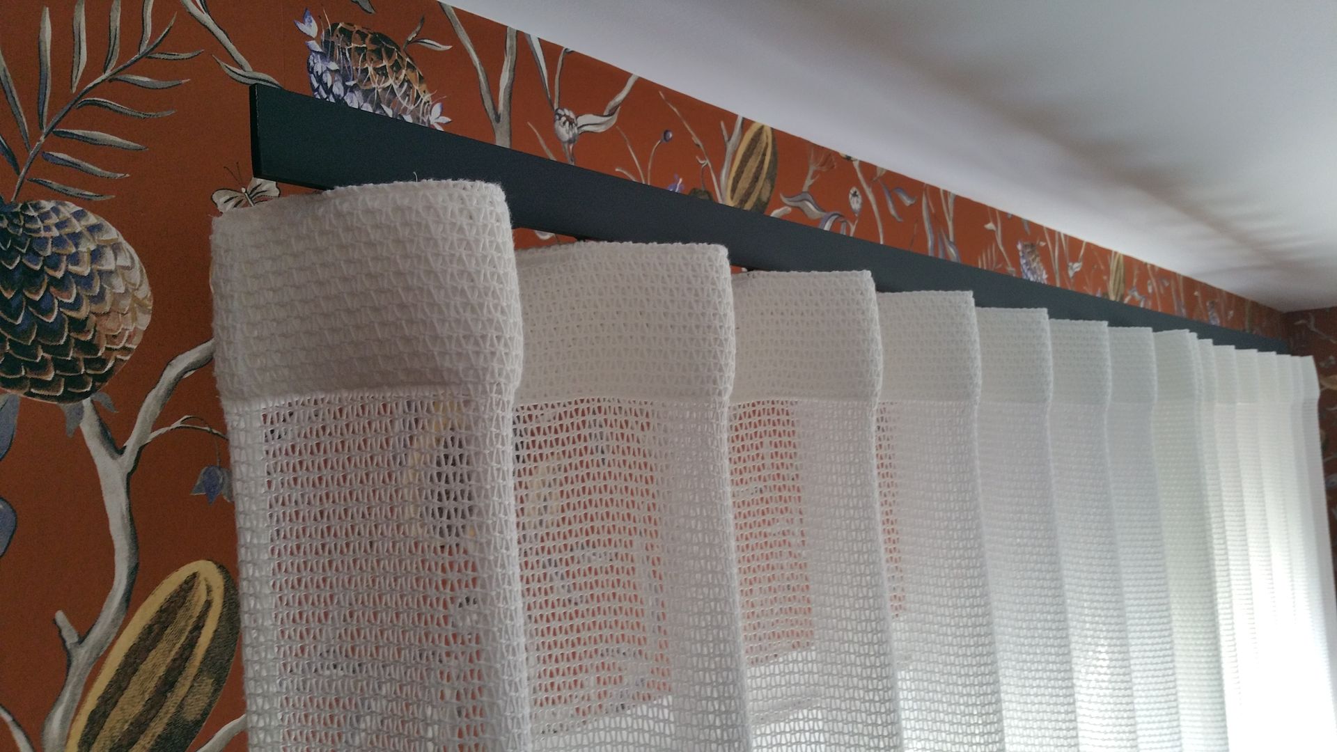 Rideau wave blanc à grandes mailles sur rail à rideau noir mat, le papier peint au mur est à fond marron avec de grand motifs rappelant un sous-bois exotique