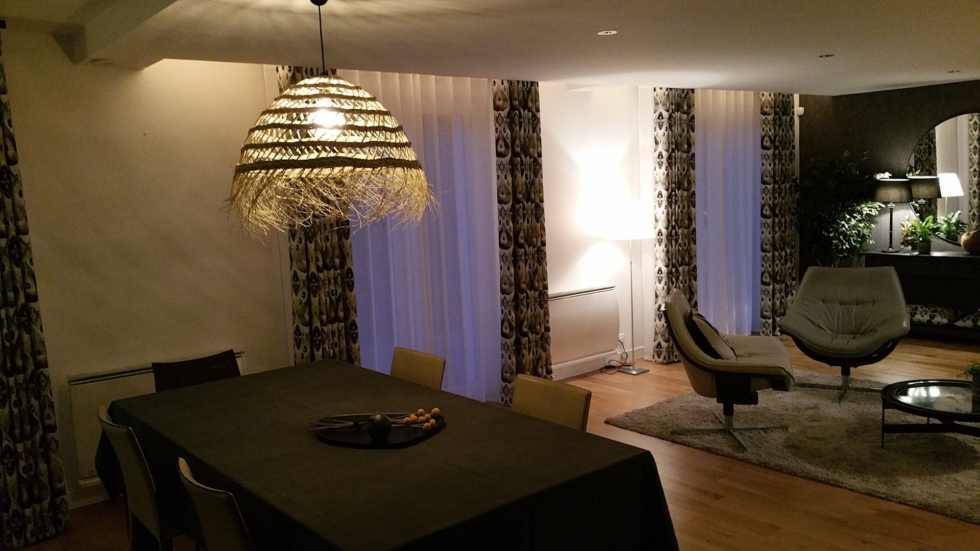 Salon avec lumière tamisée, table à manger et fauteuils pivotants. Double-rideaux et voilages devant les baies.