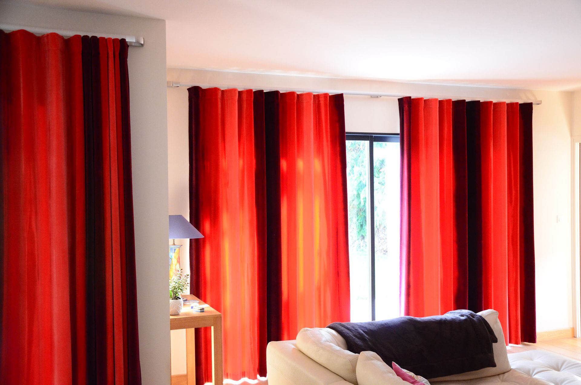 Salon avec divan blanc, petite table haute portant lampe décorative et petite plante verte, porte-fenêtres avec rideaux dont le tissu velour montre un dégradé de couleurs 