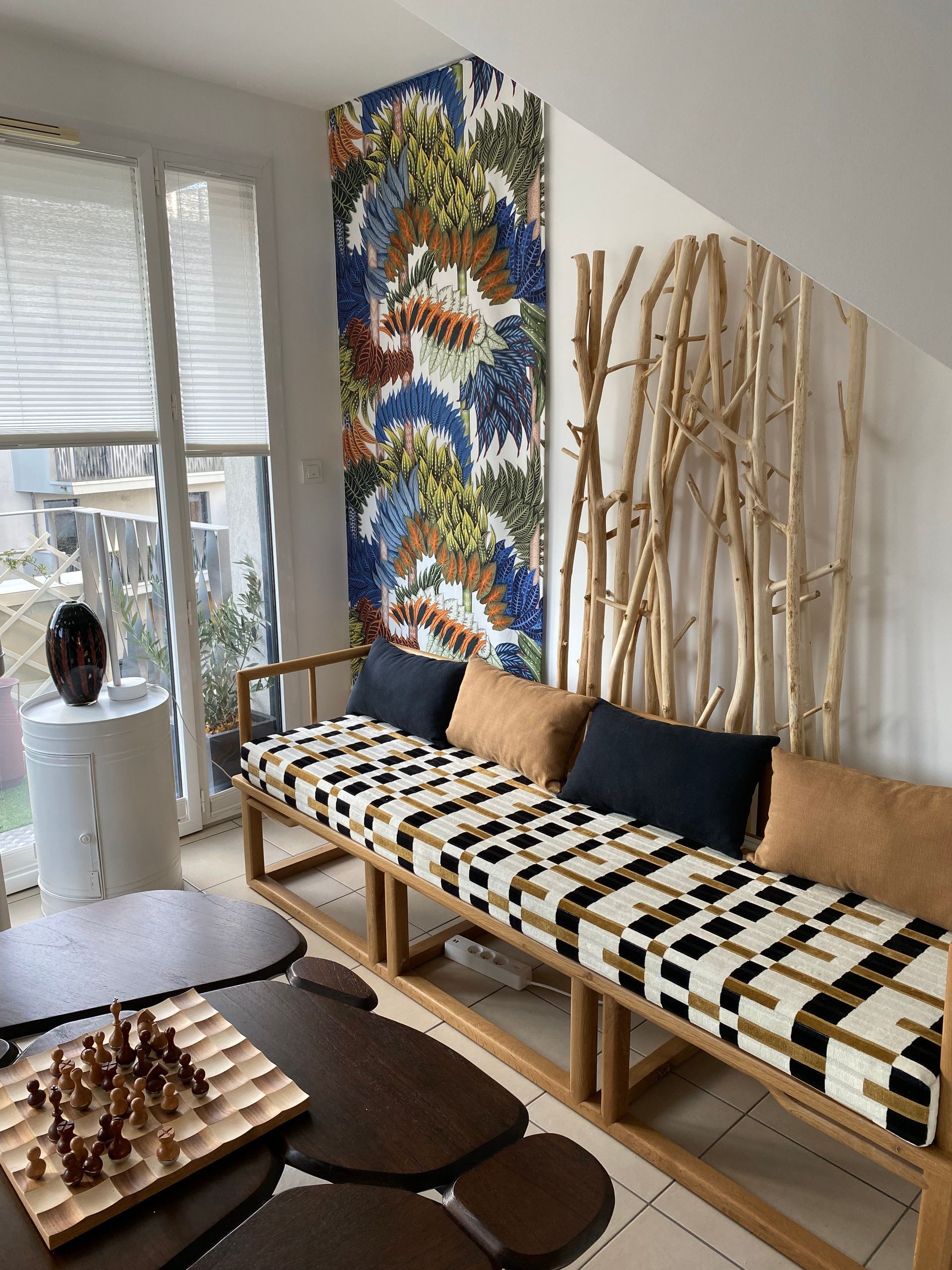 Salon avec banquette tissu moderne. Table basse et jeux d'échecs. Tenture tissu motifs végétaux très colorée au mur.. Stores plissés blancs devant les ouvertures.