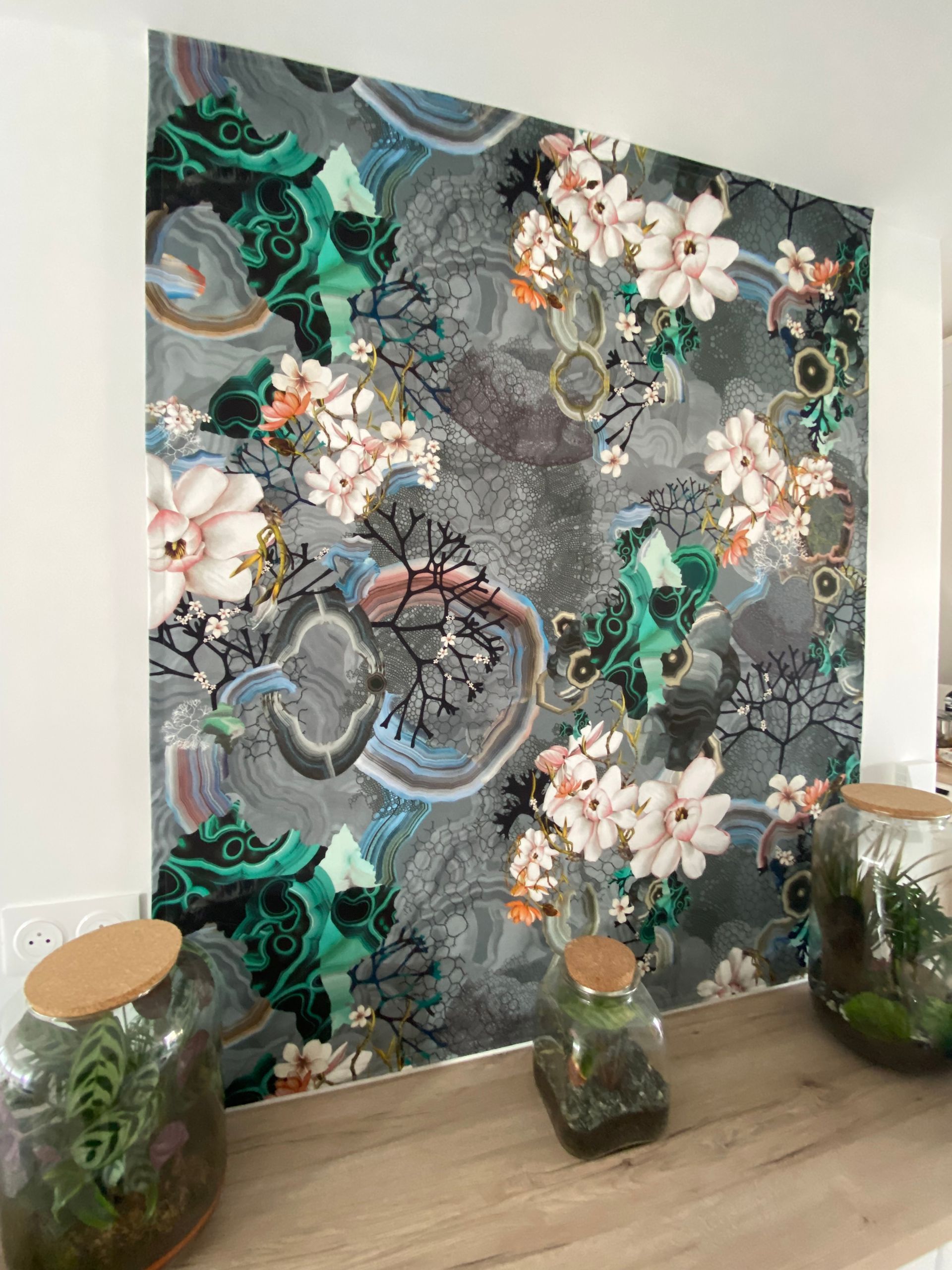 Tissu de décoration avec motifs floraux accroché à un mur comme un tableau. 3 terrariums au premier plan.