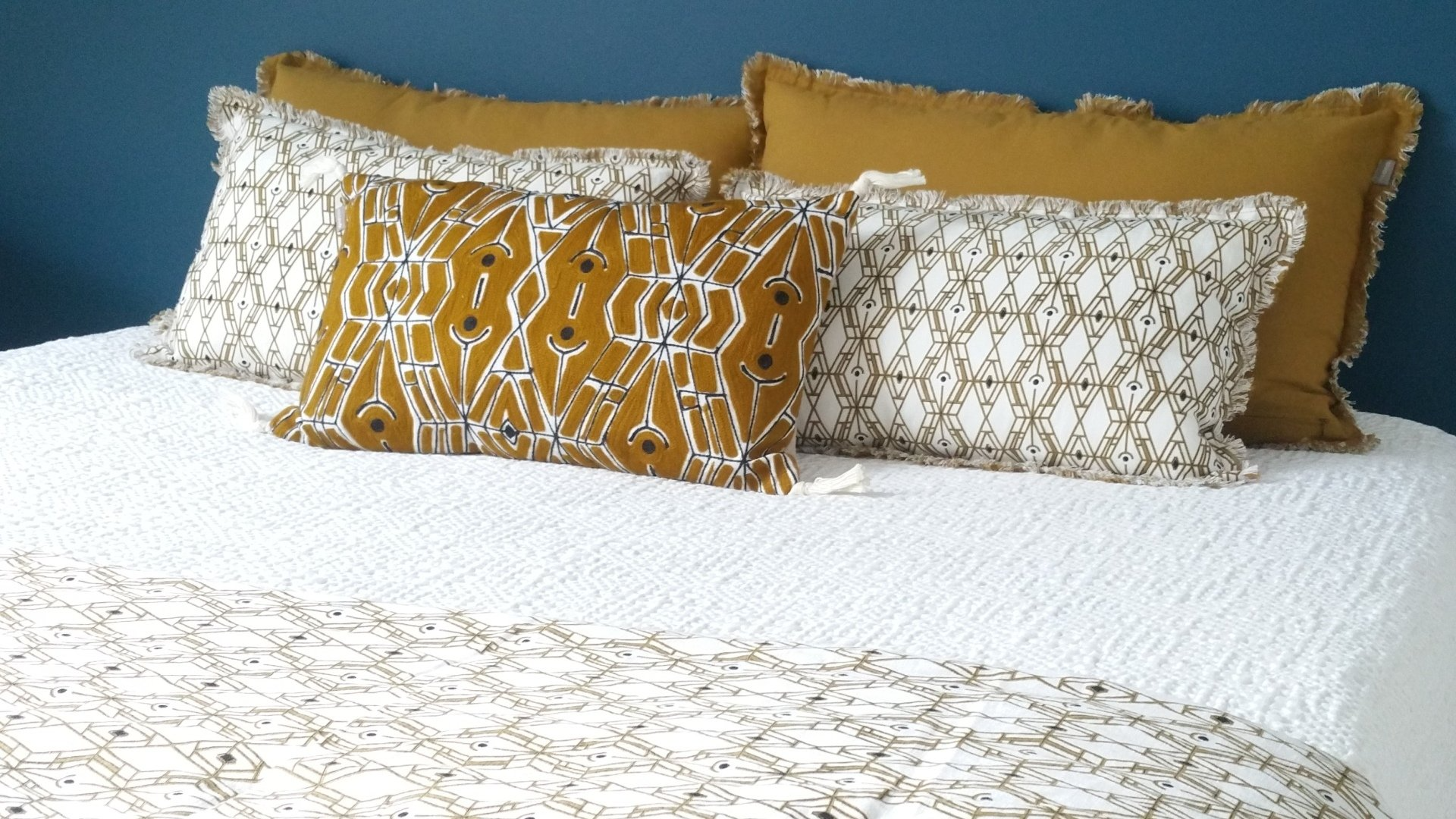 Lit orné d'un dessus de lit blanc, travers de lit ethnique et coussins de couleur blanche et/ou ocre avec motifs ethniques. Le mur derrière le lit est bleu