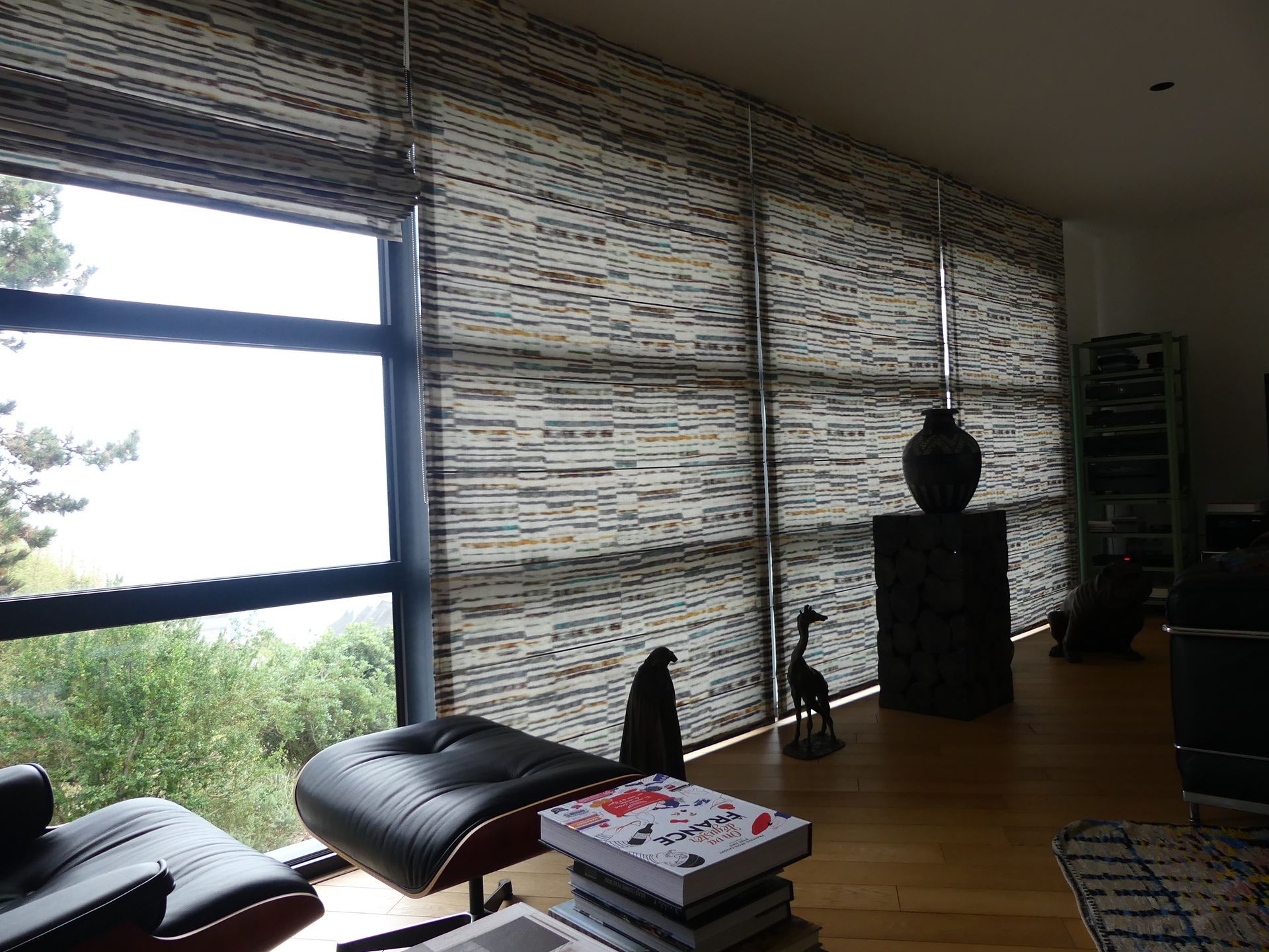 Grand salon avec fauteuil et footstep cuirs au premiers plan, puis devant baie vitrée une grande enfilade de stores bateaux allant du sol au plafond, le tissu utilisé est zébré de points et de traits horizontalement