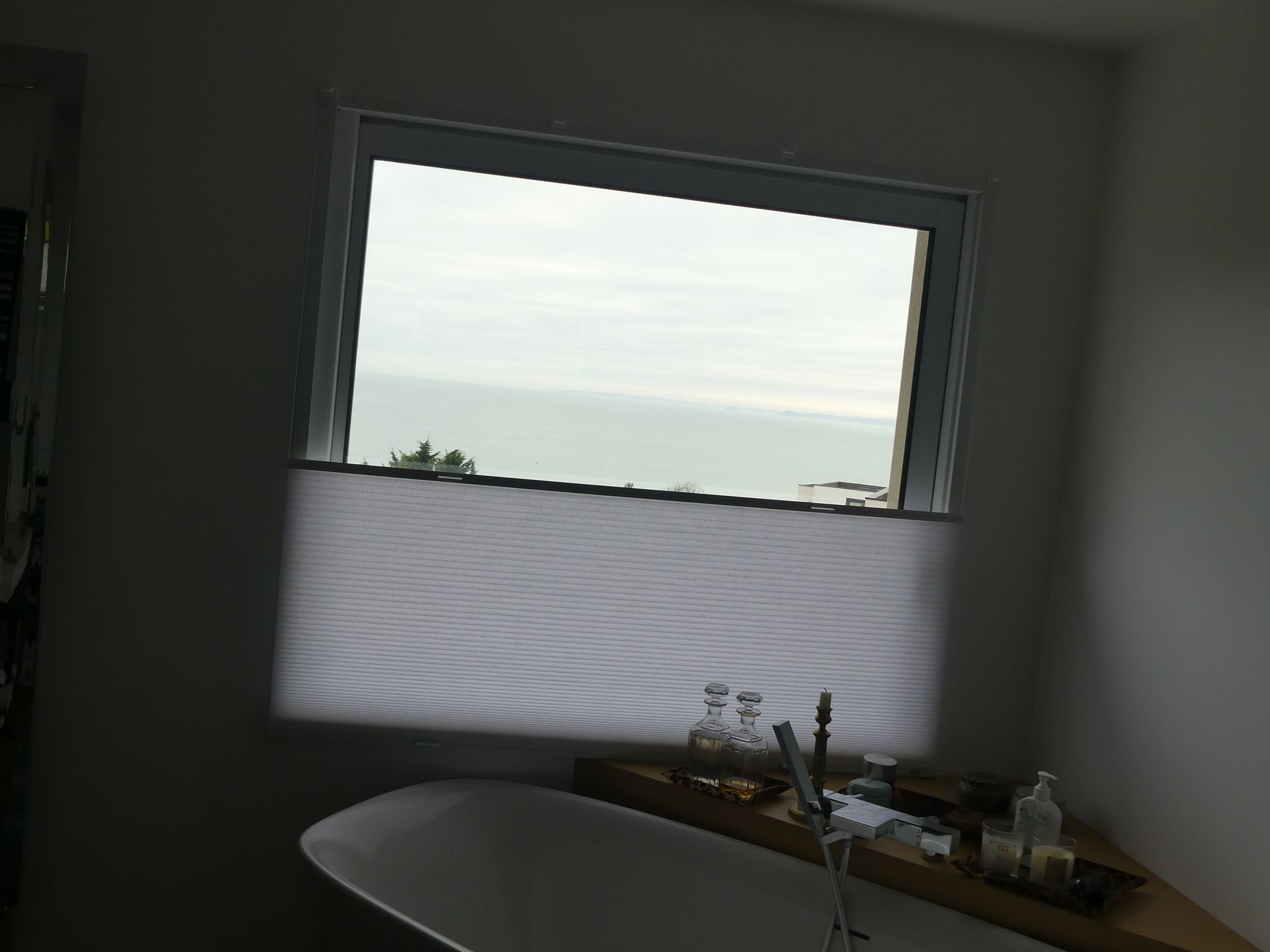 Salle de bains avec baignoire devant la baie vitrée, store plissé blanc positionné en partie basse de la vitre pour garantir l'intimité