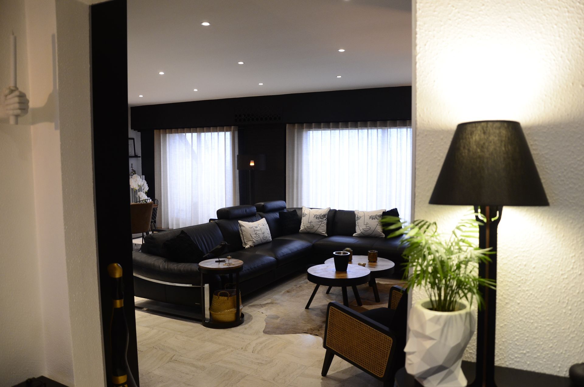 Entrée d'un salon avec canapé d'angle bleu marine et coussins blancs. Voilages clairs façon rideaux waves devant les baies.