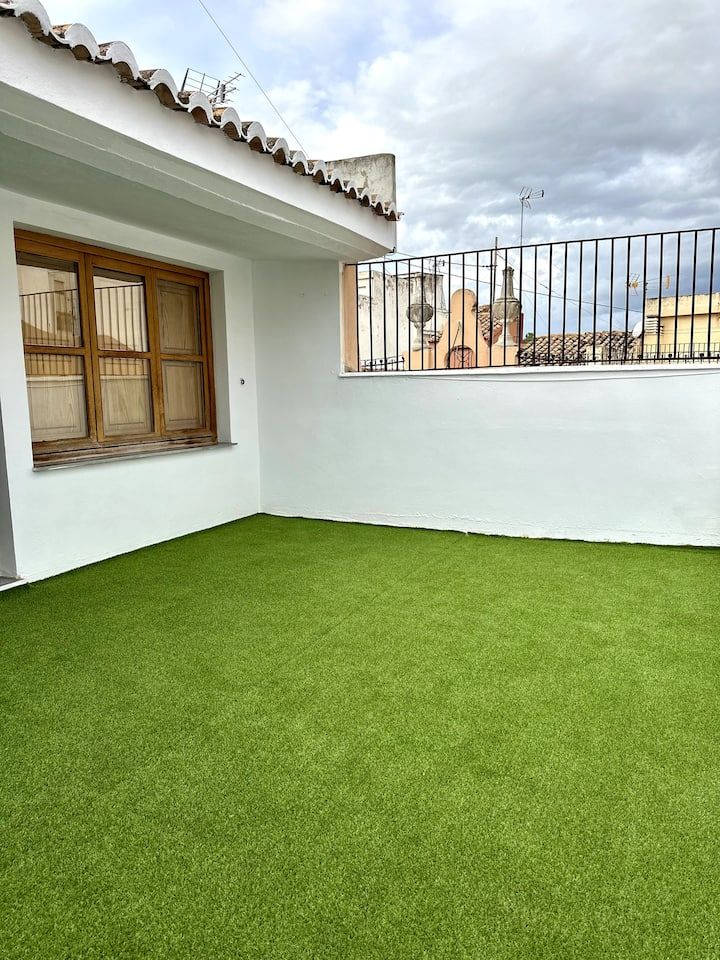 Pequeño patio en la azotea con césped verde, paredes blancas y barandilla de hierro forjado. Cielo nublado.