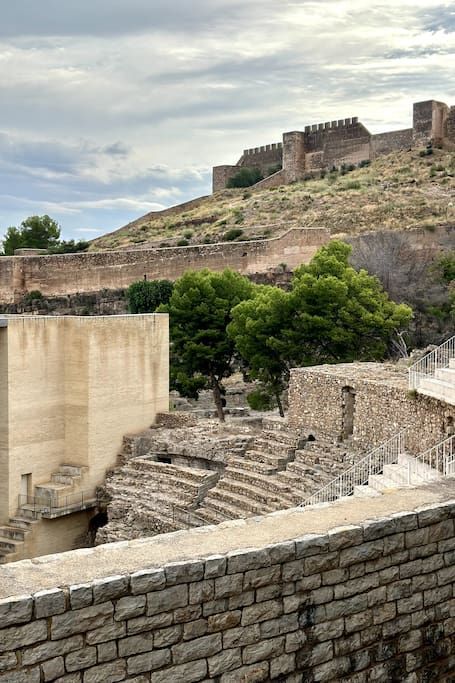 Antiguo castillo de piedra en lo alto de una colina, con ruinas y escaleras en primer plano y un cielo nublado arriba.