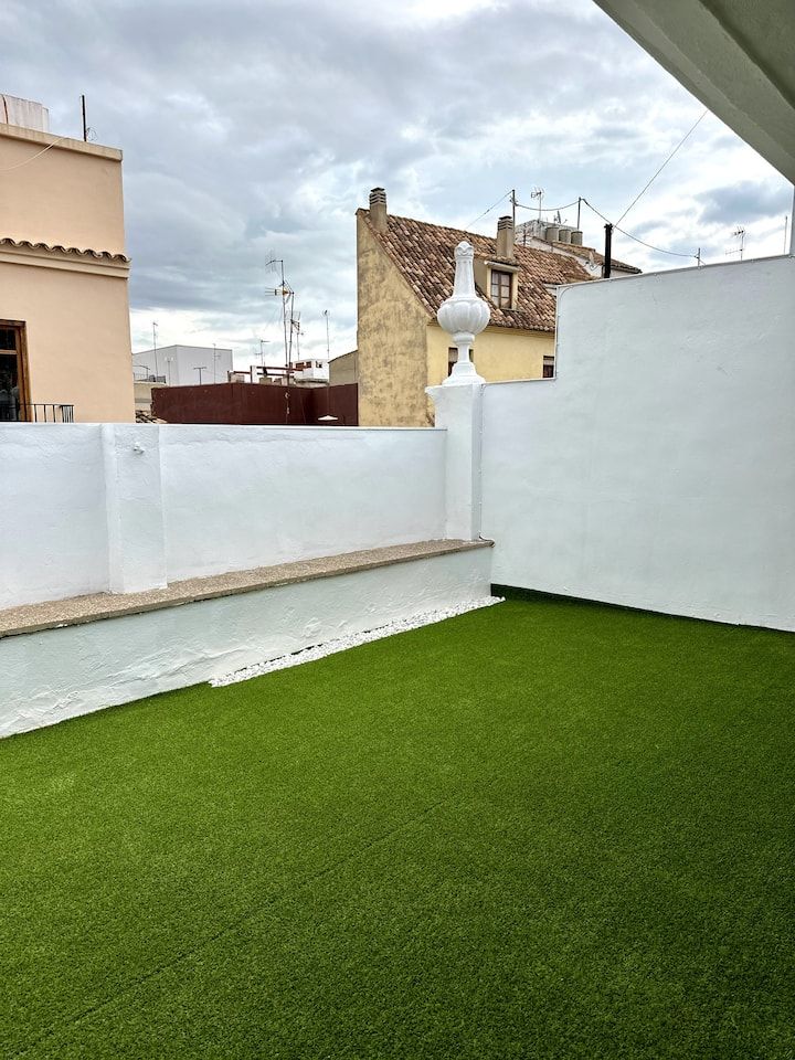 Patio en la azotea con césped artificial verde, paredes blancas y un cielo nublado.