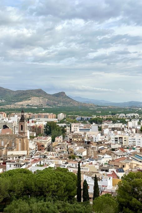 Paisaje urbano con torre de iglesia, edificios, árboles y montañas distantes bajo un cielo nublado.
