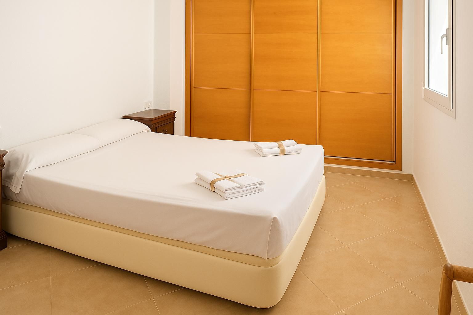 Dormitorio con cama, mesita de noche, armario de madera y una pequeña ventana.