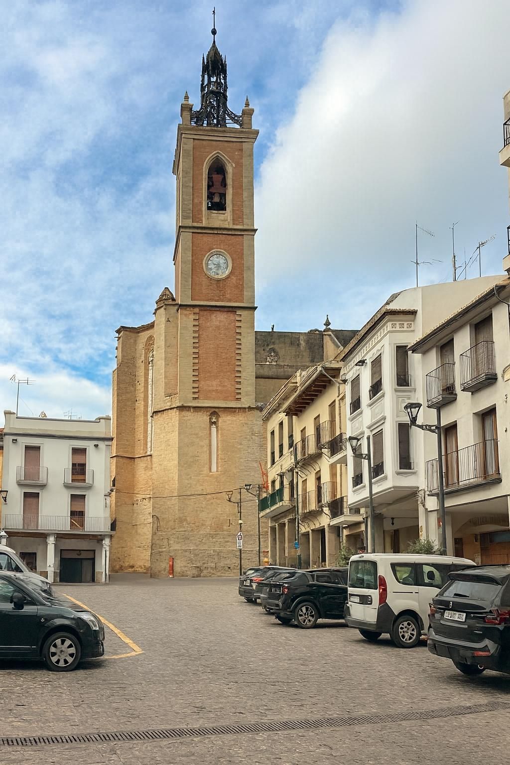 Plaza con una alta torre de iglesia de ladrillo y piedra, rodeada de edificios y coches aparcados. Cielo azul con nubes.