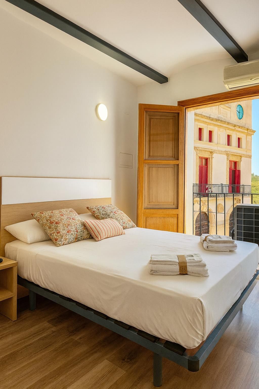 Dormitorio con cama blanca, puerta de madera abierta al balcón, vista exterior del edificio tapizado en rojo.