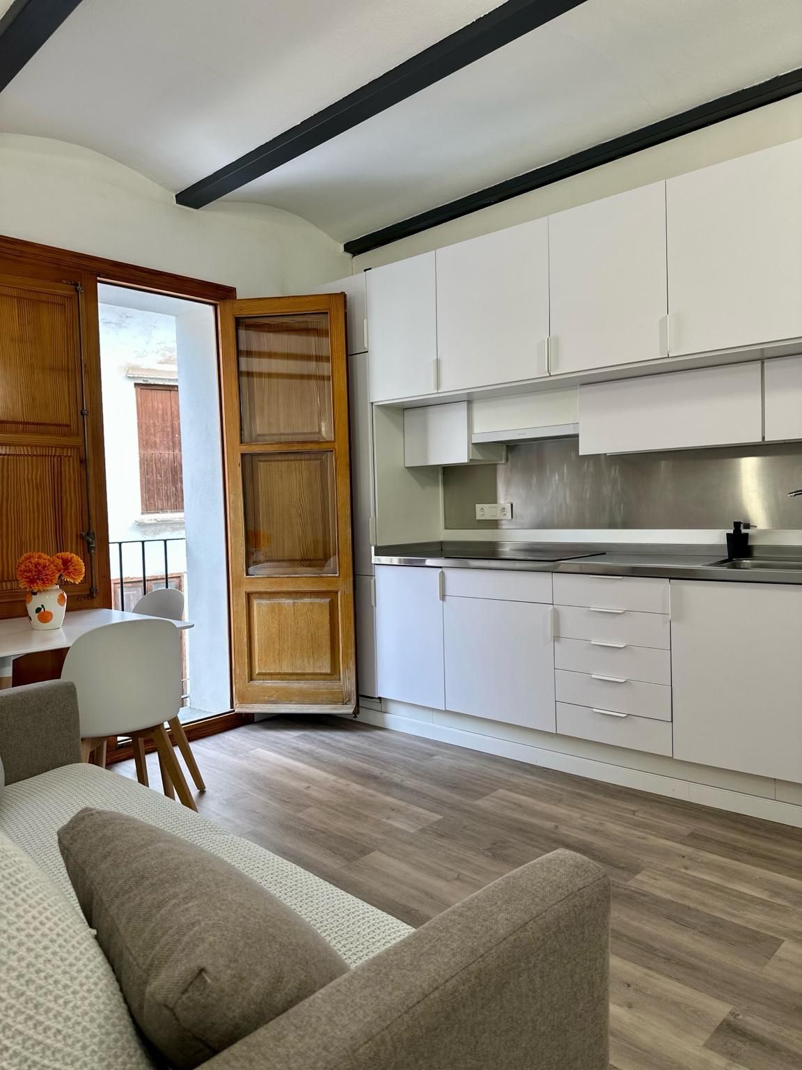 Cocina pequeña con gabinetes blancos, piso de madera y acceso a un balcón. Un sofá se encuentra en primer plano.