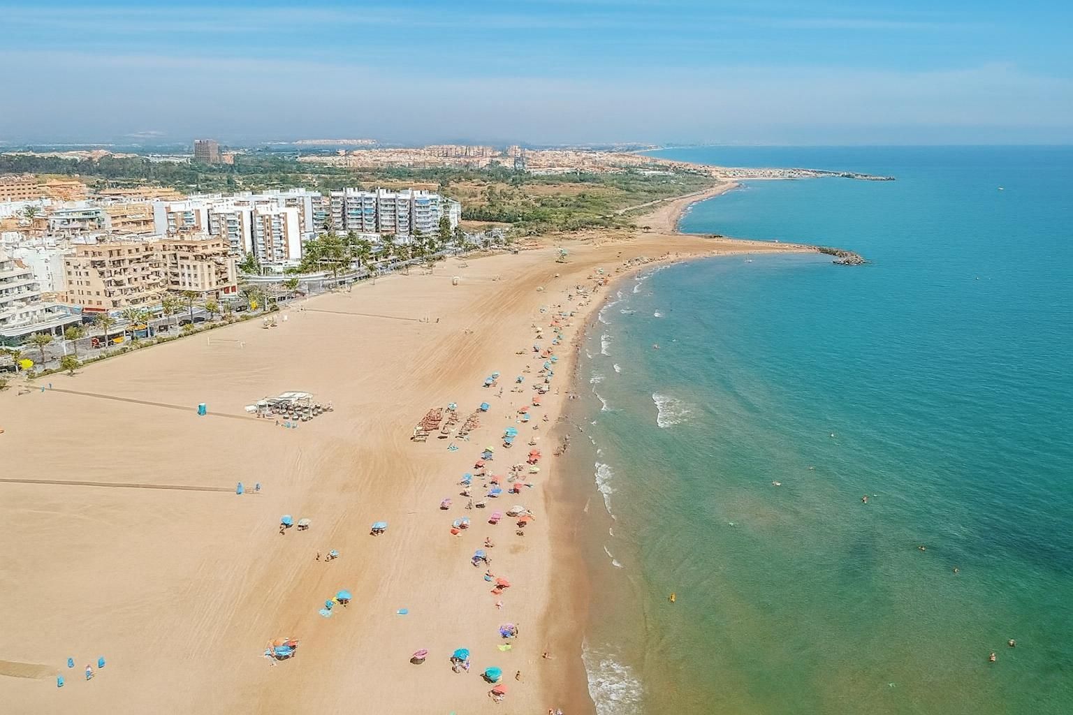Playa de arena con gente, agua turquesa y edificios a lo largo de la costa bajo un cielo azul.