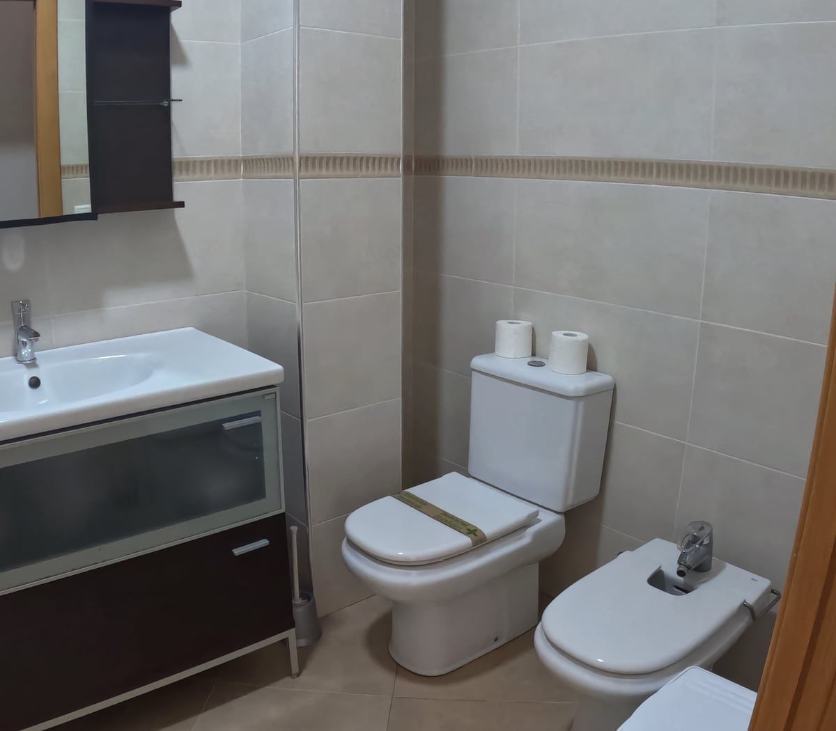 Baño con lavabo, inodoro, bidé y mueble de baño marrón. Azulejos de color claro.