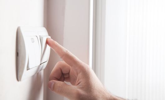 Main appuyant sur un thermostat mural blanc près d'une fenêtre aux rideaux transparents.