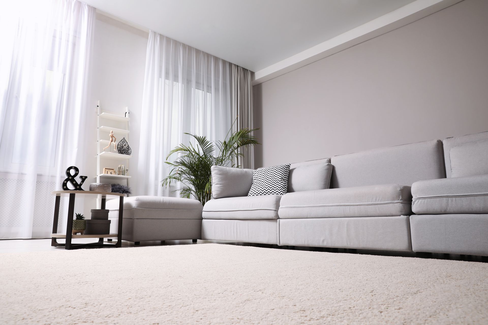 Salon lumineux avec un canapé gris, des voilages, une table d'appoint et un tapis clair.