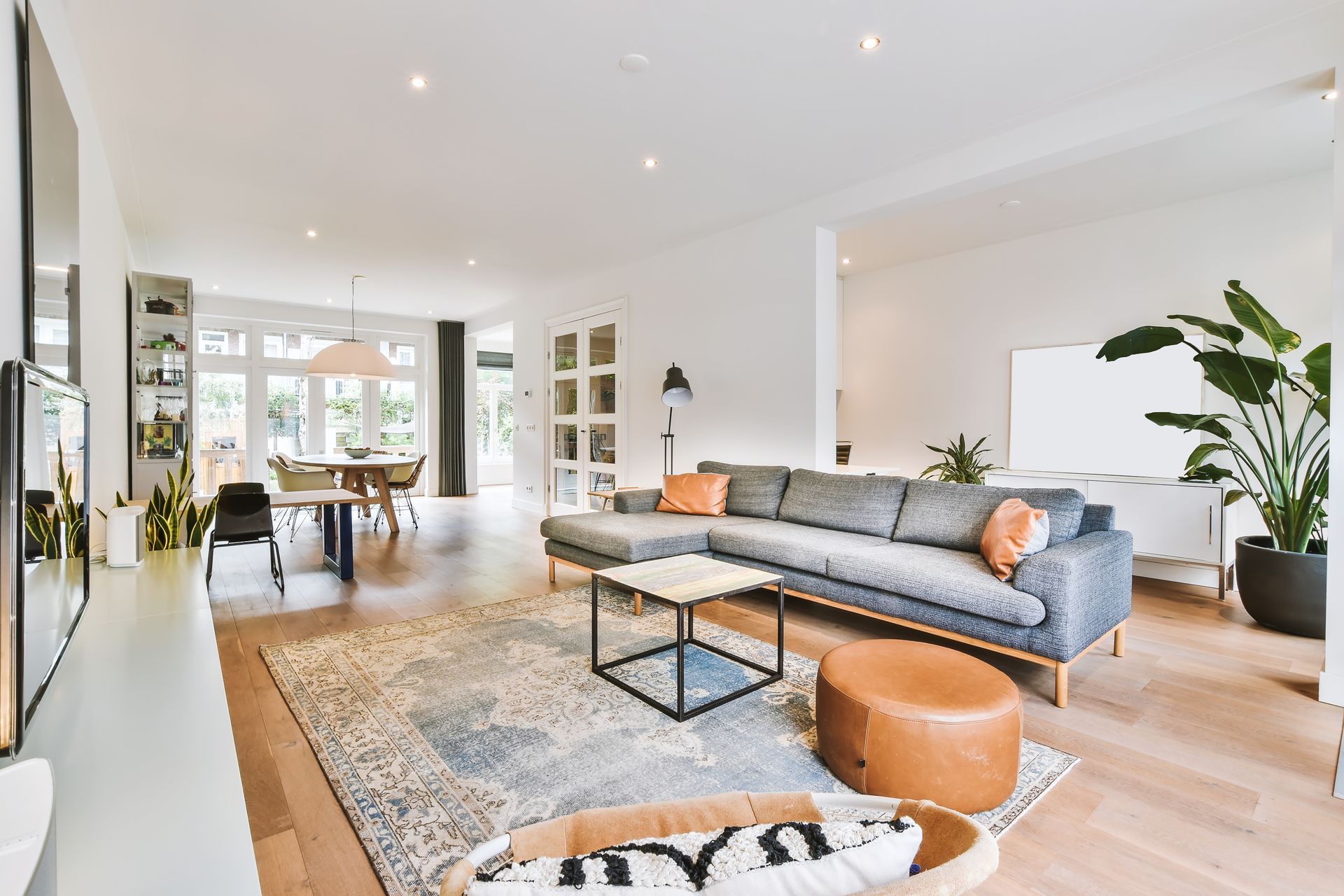 Salon lumineux et ouvert avec canapé gris, pouf orange, coin repas et grandes fenêtres.