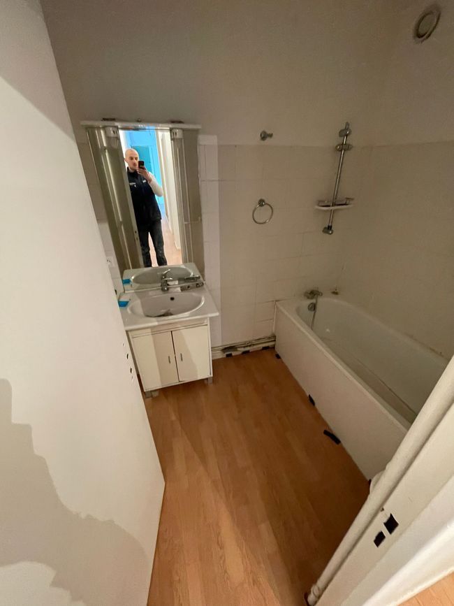 Salle de bains étroite avec baignoire, lavabo, miroir et sol imitation bois ; reflet d'une personne dans le miroir.
