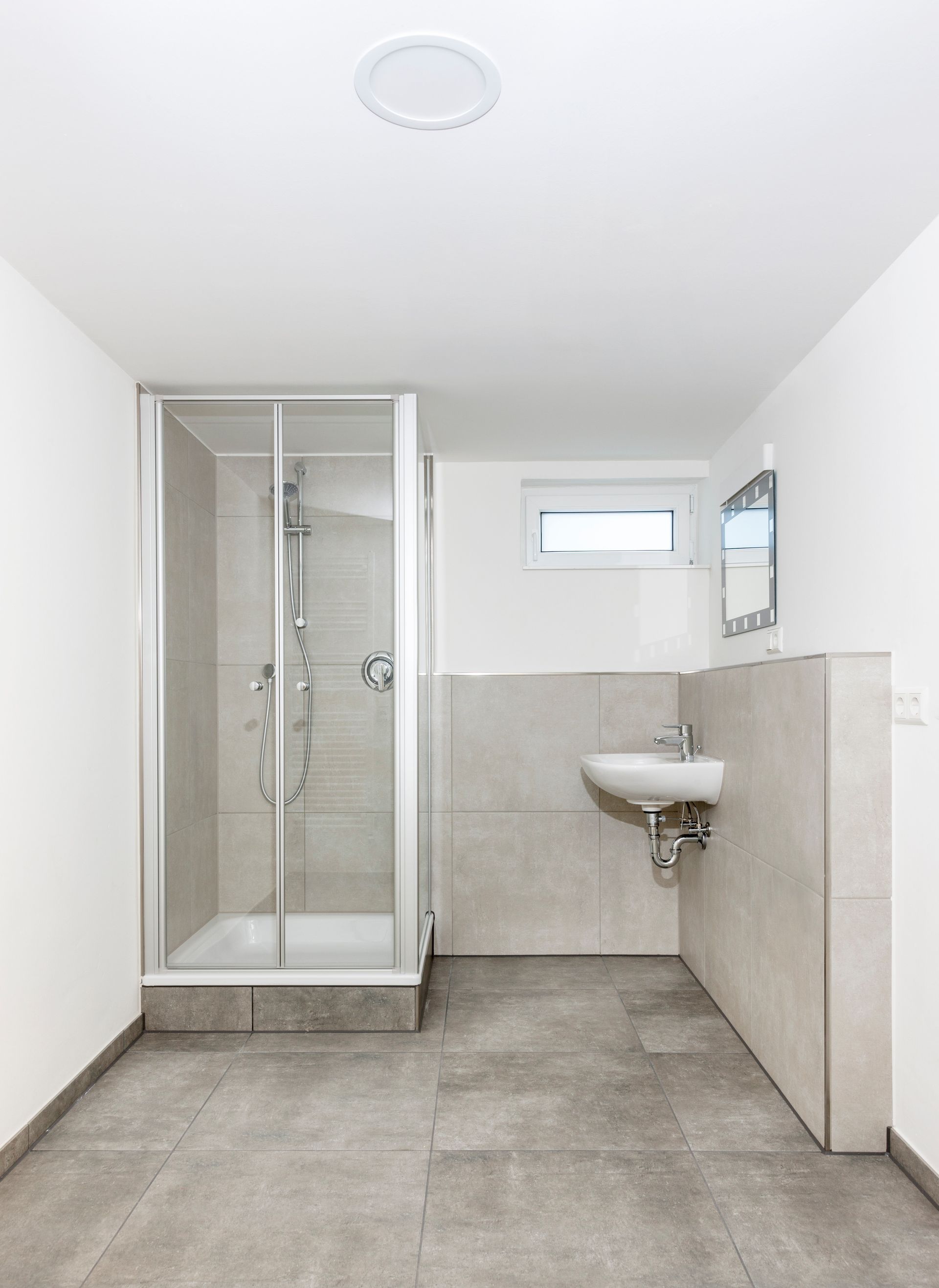 Salle de bain moderne vide avec douche en verre, lavabo suspendu, murs blancs et sol carrelé gris.
