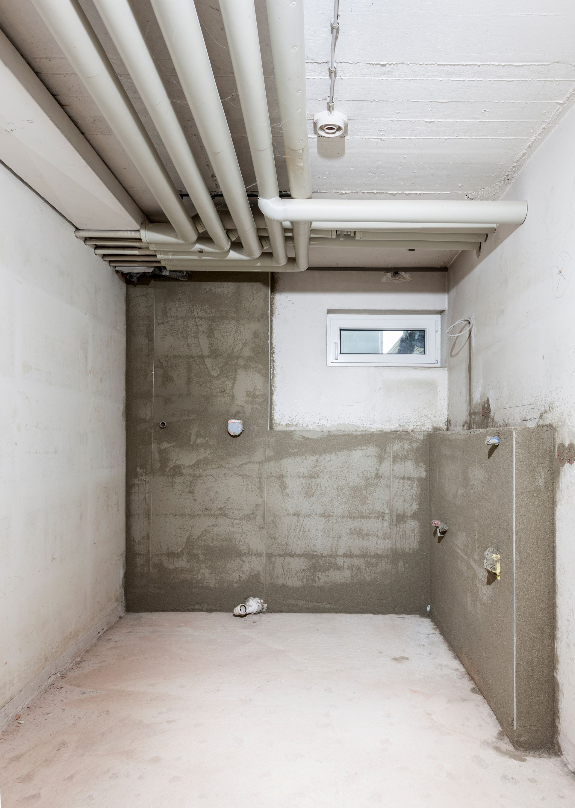 Sous-sol en béton brut, non aménagé, avec tuyaux apparents et une petite fenêtre.