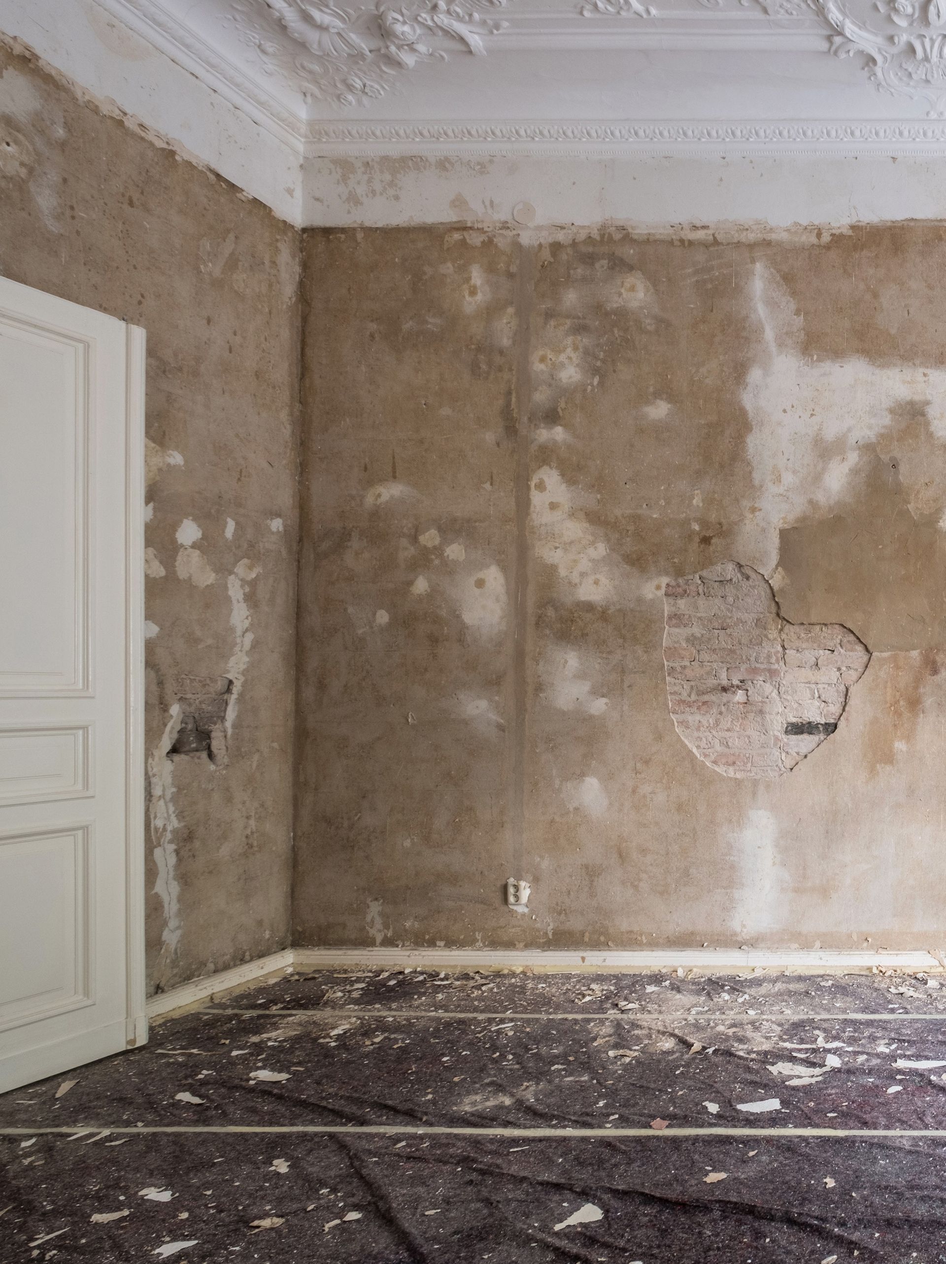Pièce abandonnée aux murs décrépis, moulures blanches au plafond et débris au sol.