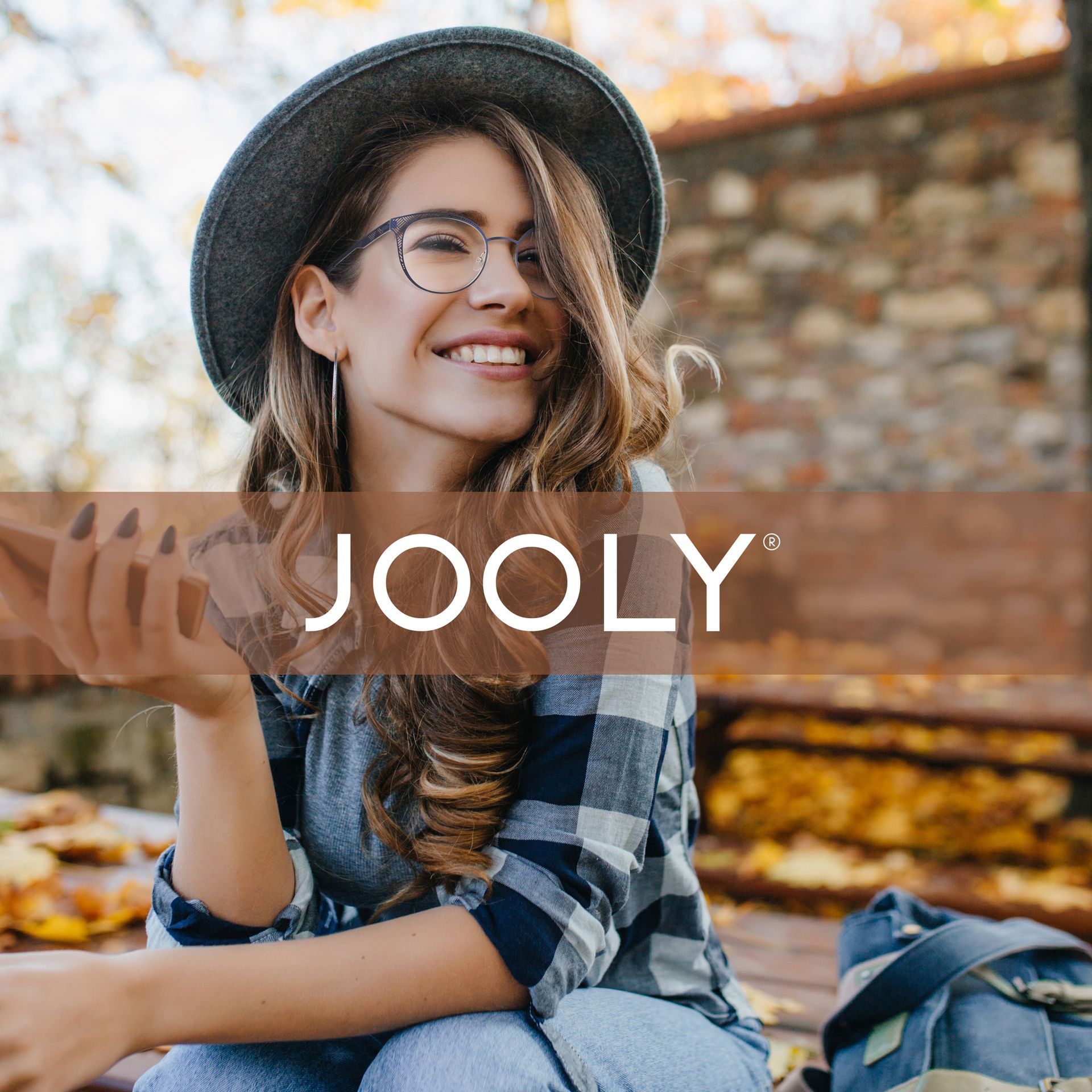 Une femme portant un chapeau et des lunettes sourit assise sur un banc.