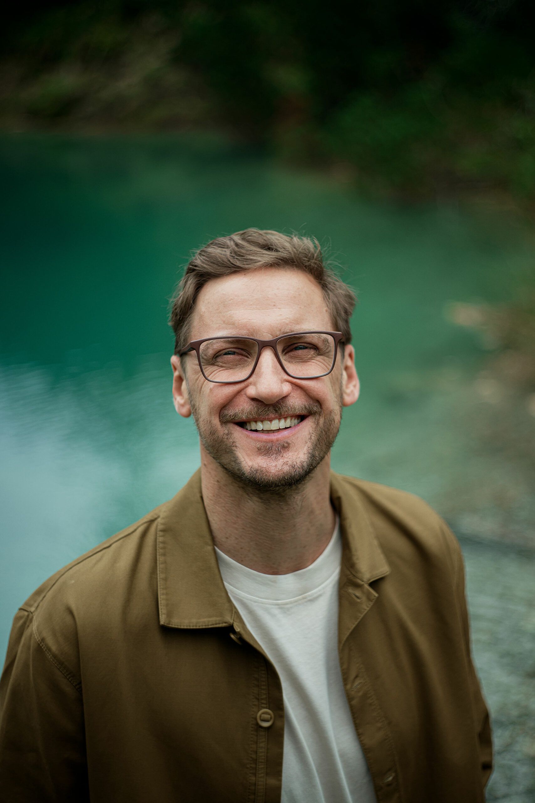 Un homme portant des lunettes et une veste marron sourit devant un lac.