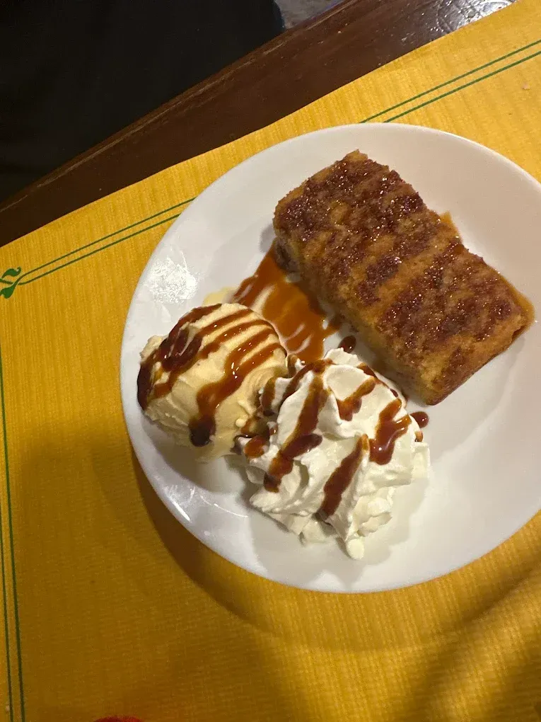 Plato blanco con un postre bañado en caramelo y un pastel rectangular glaseado sobre un mantel individual amarillo.