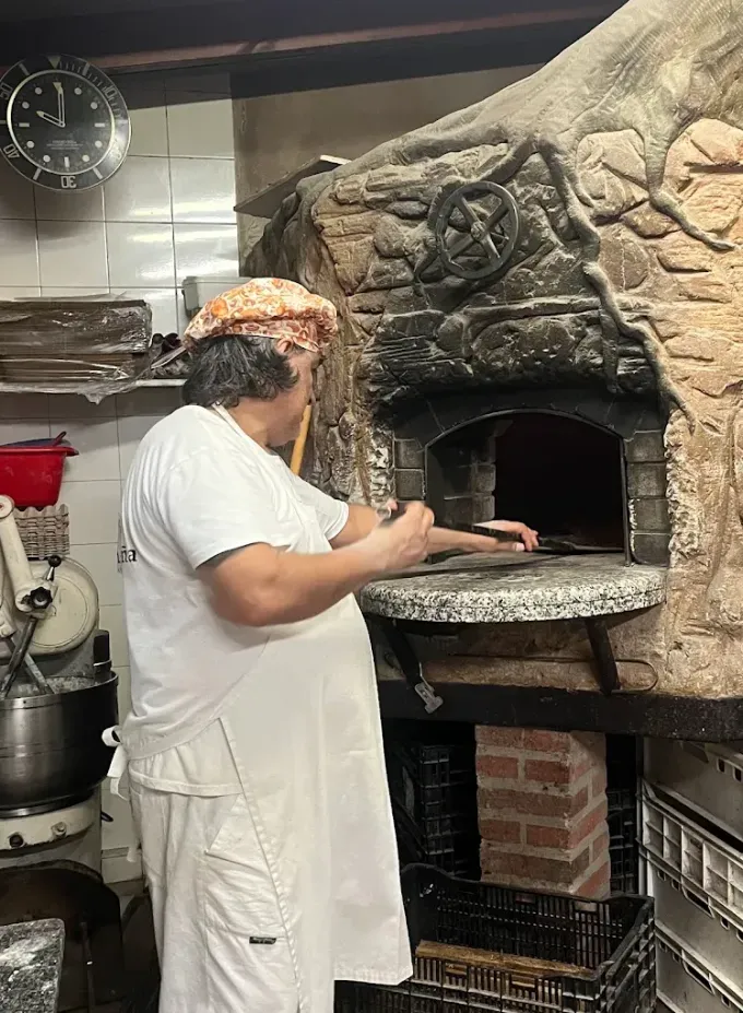 Una persona con delantal blanco coloca una pizza en un horno de piedra en una cocina.