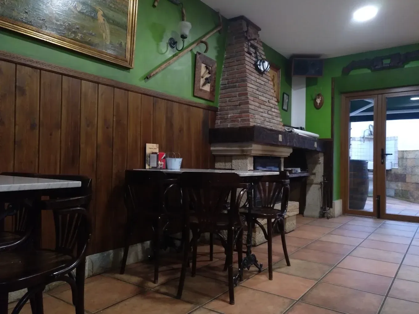 Interior de cafetería rústica vacía con paredes verdes, revestimiento de madera, chimenea de ladrillo y suelo de baldosas.