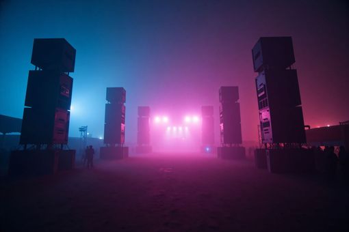 Scène de concert enveloppée de brume, baignées par des néons roses et bleus, avec de hautes tours d'enceintes.