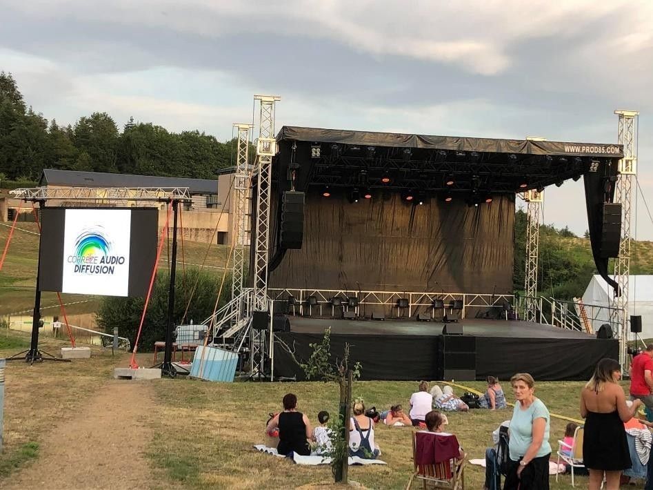 Scène de concert en plein air, avec une pancarte annonçant un festival et quelques personnes assises sur la pelouse.