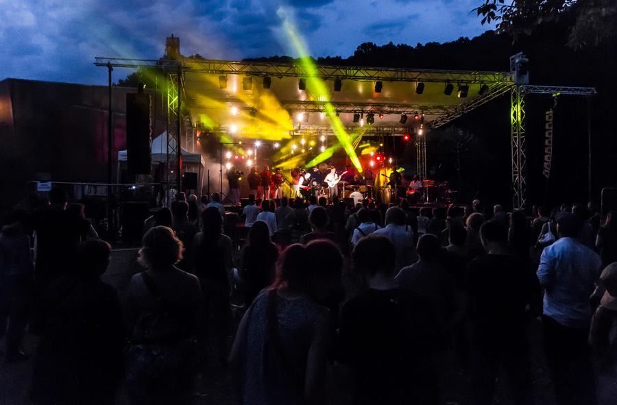 Scène de concert en plein air, avec une pancarte annonçant un festival et quelques personnes assises sur la pelouse.