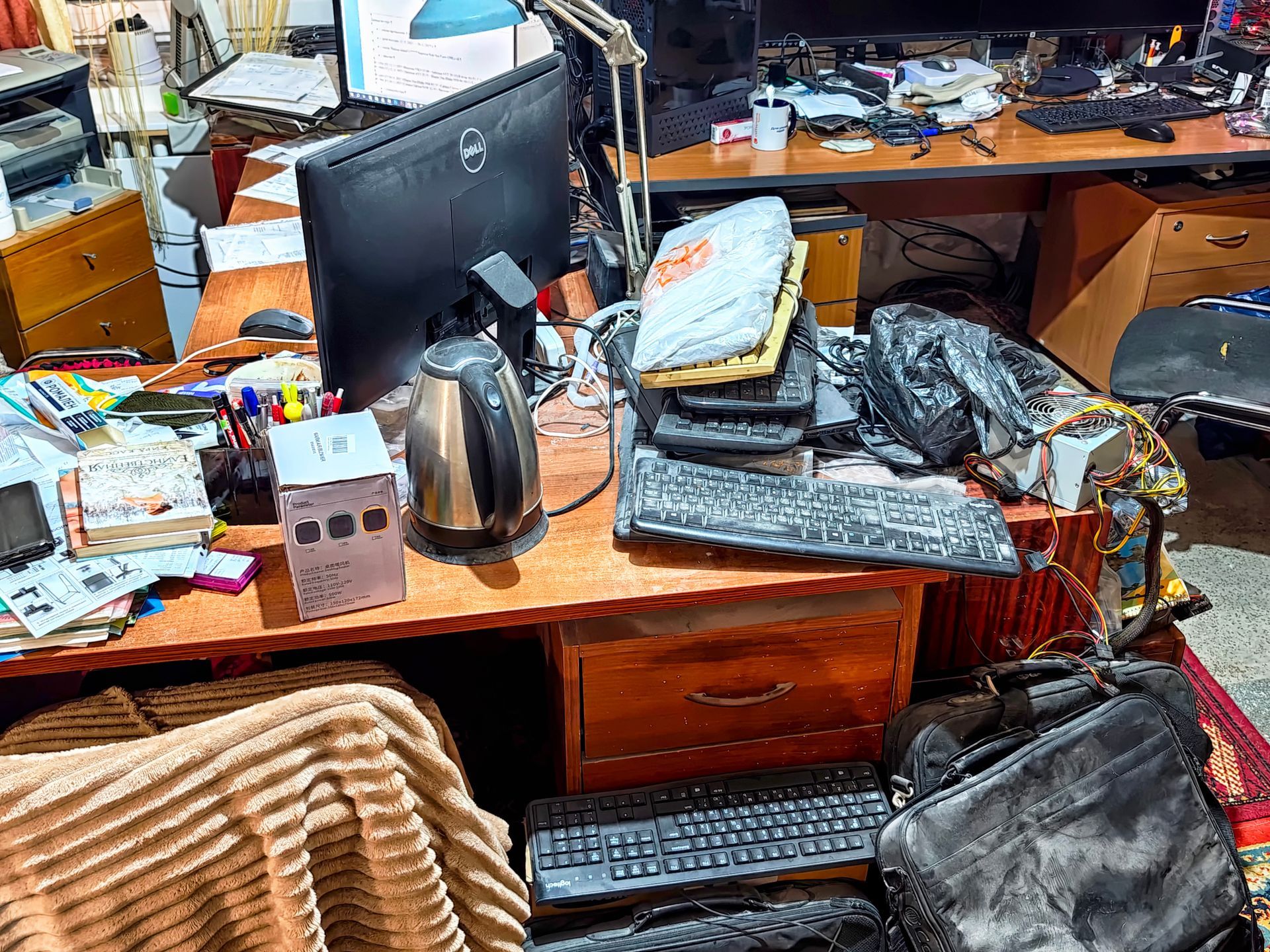 Bureau encombré avec écran d'ordinateur, clavier, bouilloire, papiers et sacs empilés autour.
