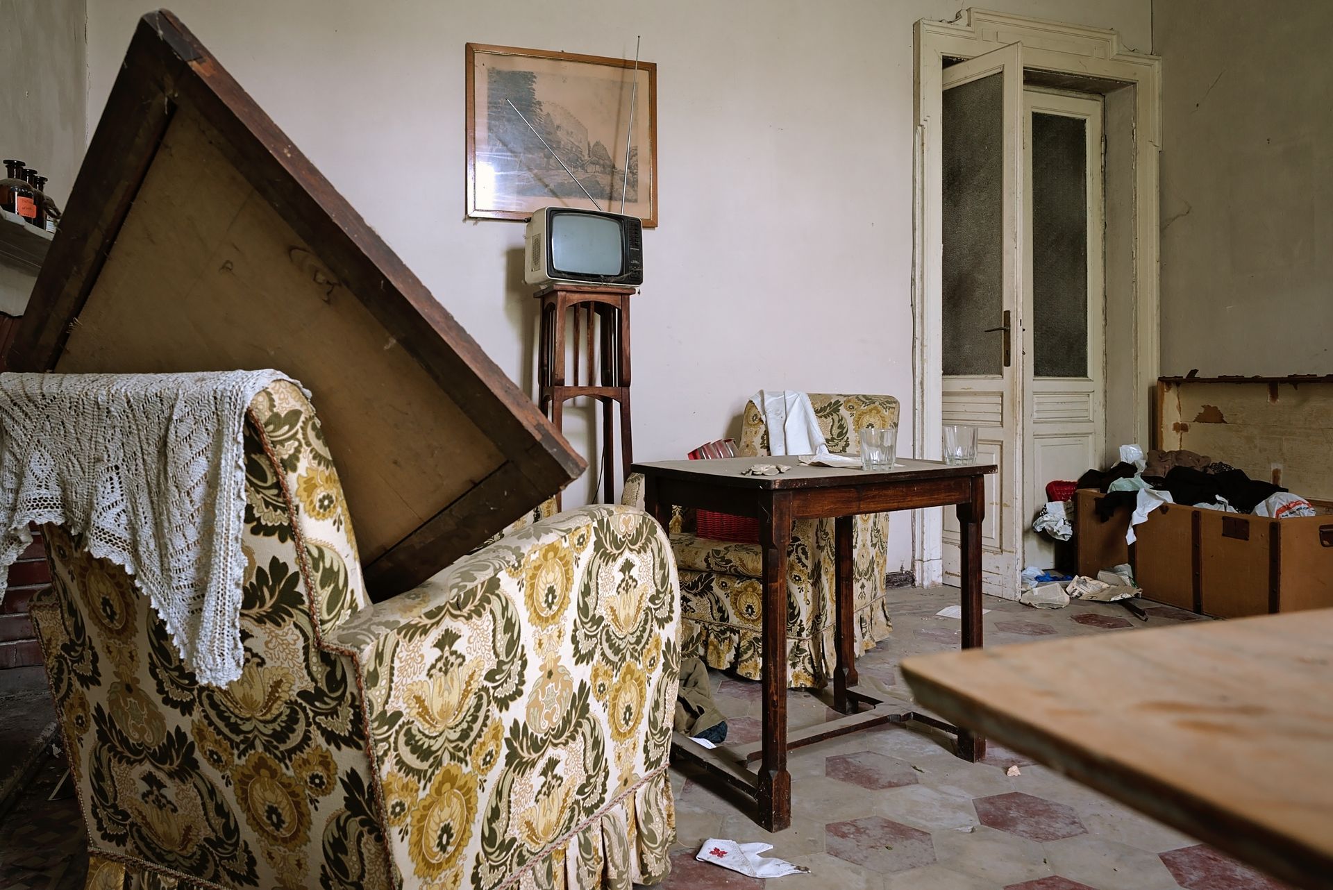 Une pièce abandonnée avec un fauteuil à motifs floraux, une petite table en bois, une télévision vintage et des cartons encombrés.