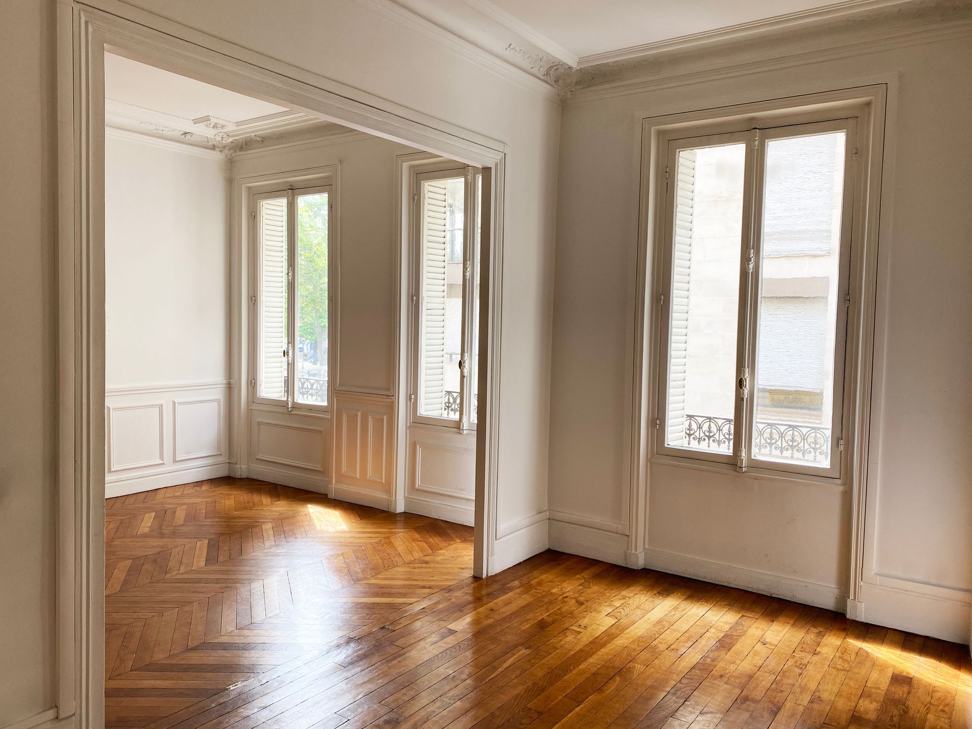 Pièce lumineuse et vide avec parquet à chevrons, murs blancs, moulures décoratives et grandes fenêtres ensoleillées.