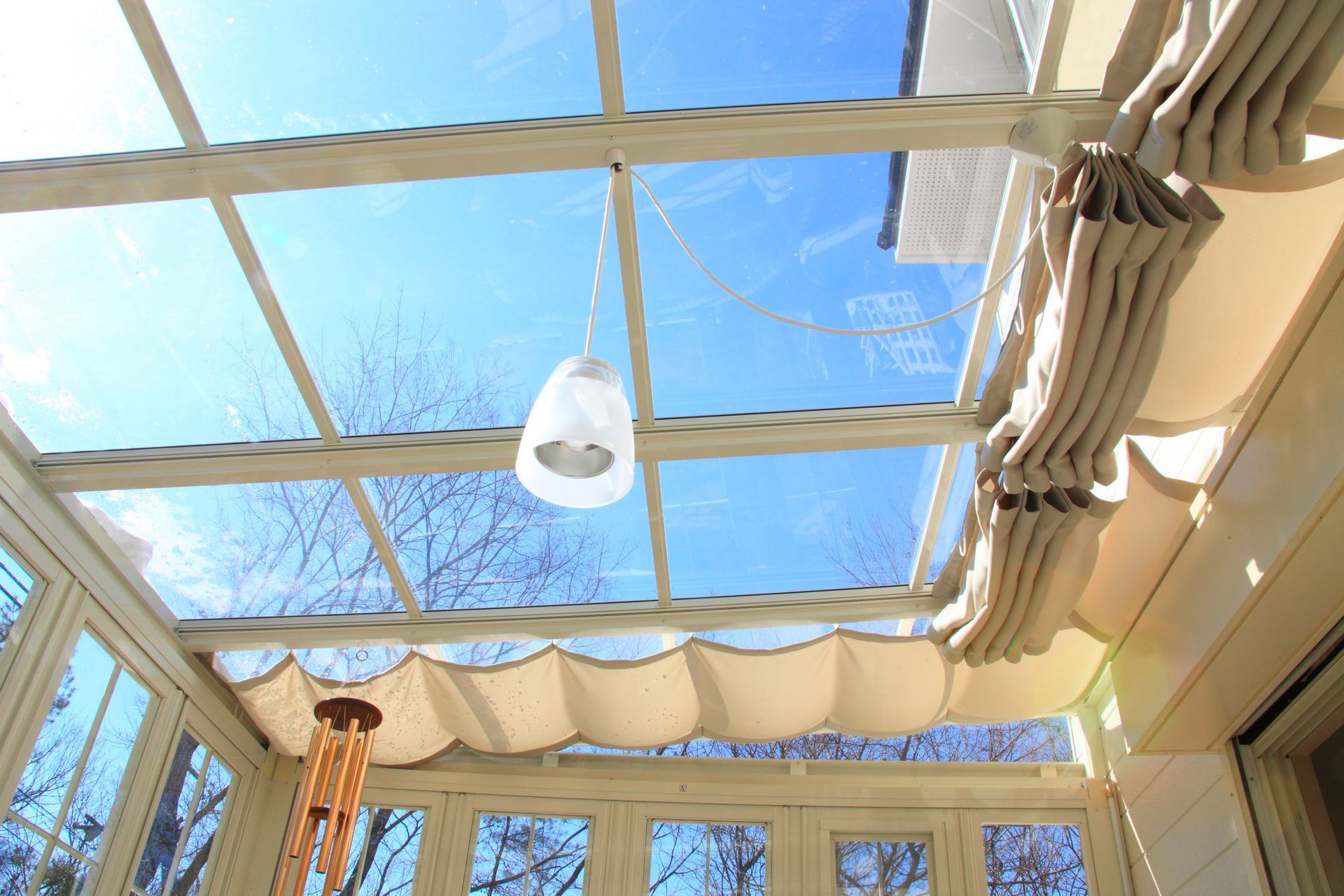 Plafond vitré ensoleillé laissant voir le ciel bleu, encadré de rideaux beiges et d’un luminaire suspendu