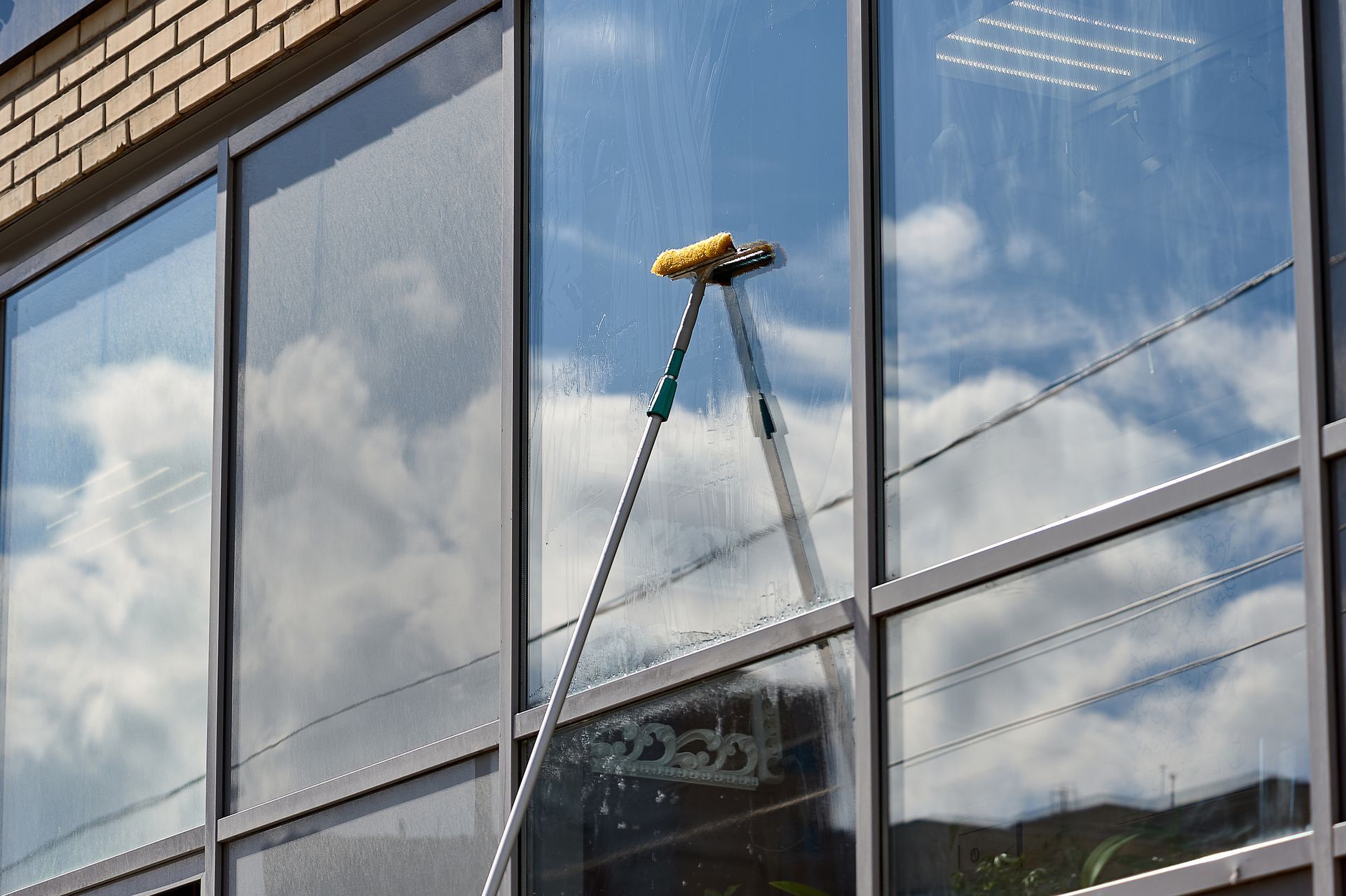 Un laveur de vitres nettoie la façade vitrée d’un immeuble à l’aide d’une longue raclette, qui reflète les nuages et le ciel