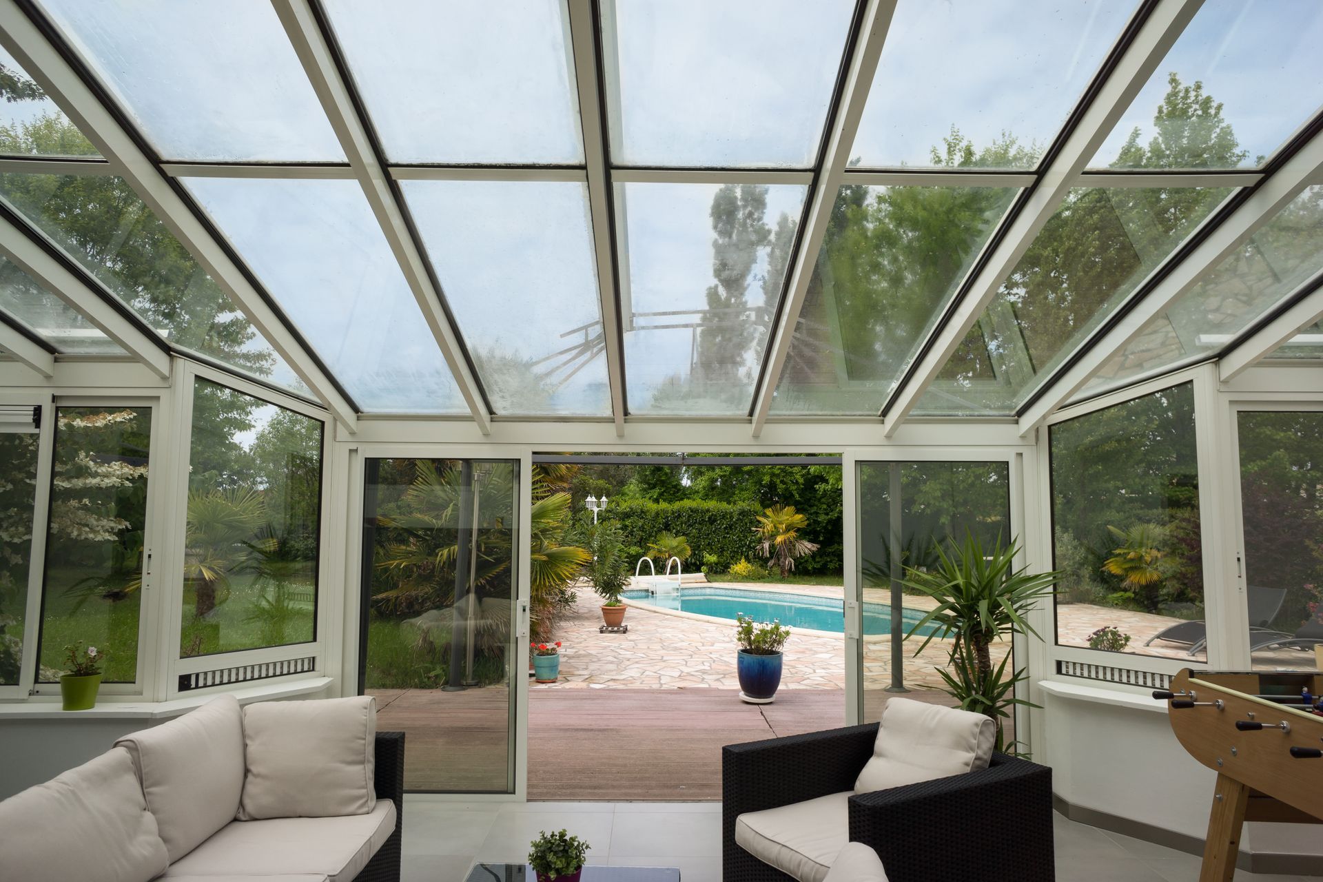 Véranda vitrée avec un canapé blanc, un fauteuil sombre et des portes coulissantes donnant sur une piscine et une terrasse dans le jardin.