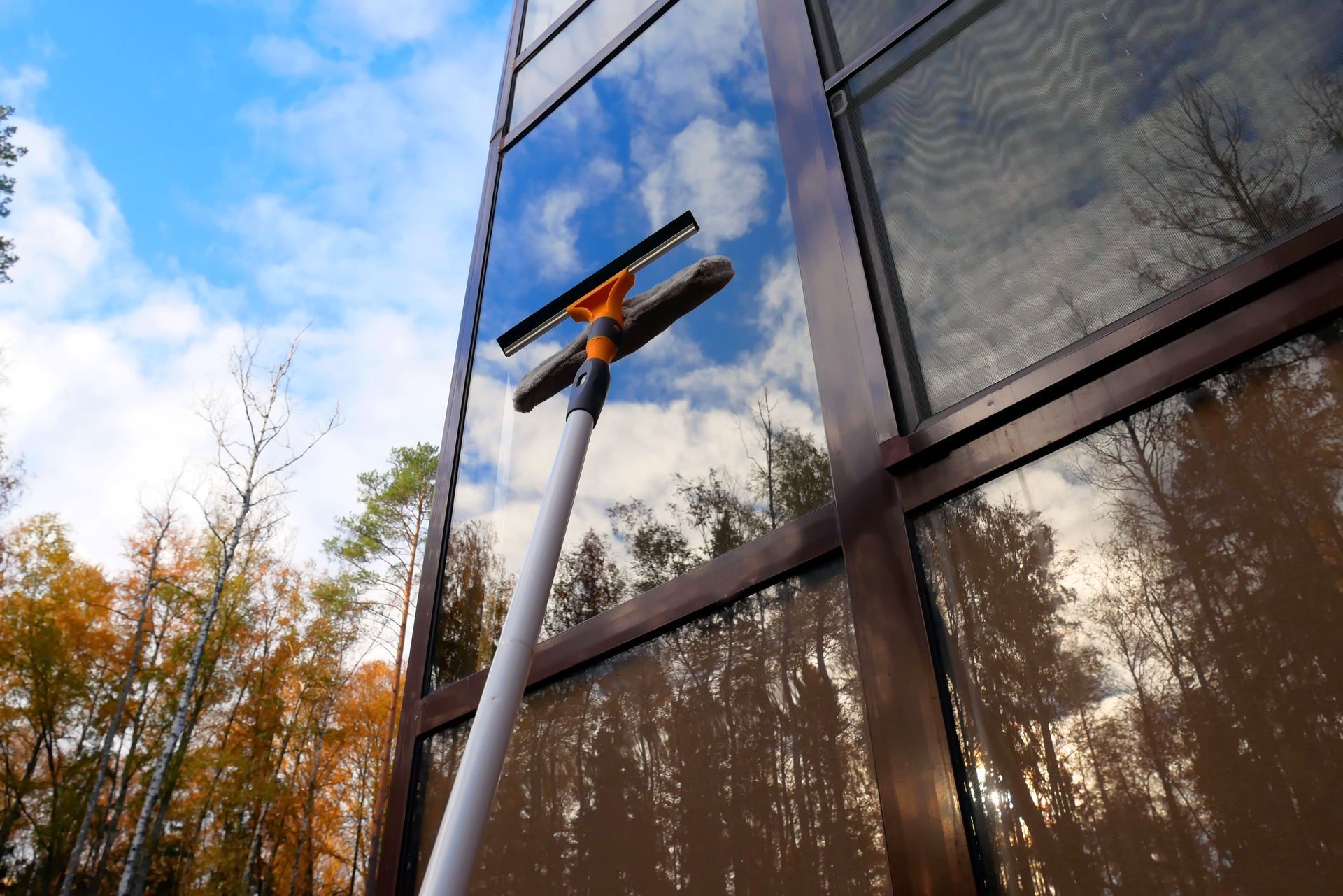 Outil de nettoyage des vitres sur la façade d’un bâtiment, sous un ciel bleu, avec des arbres d’automne qui se reflètent dans les vitres