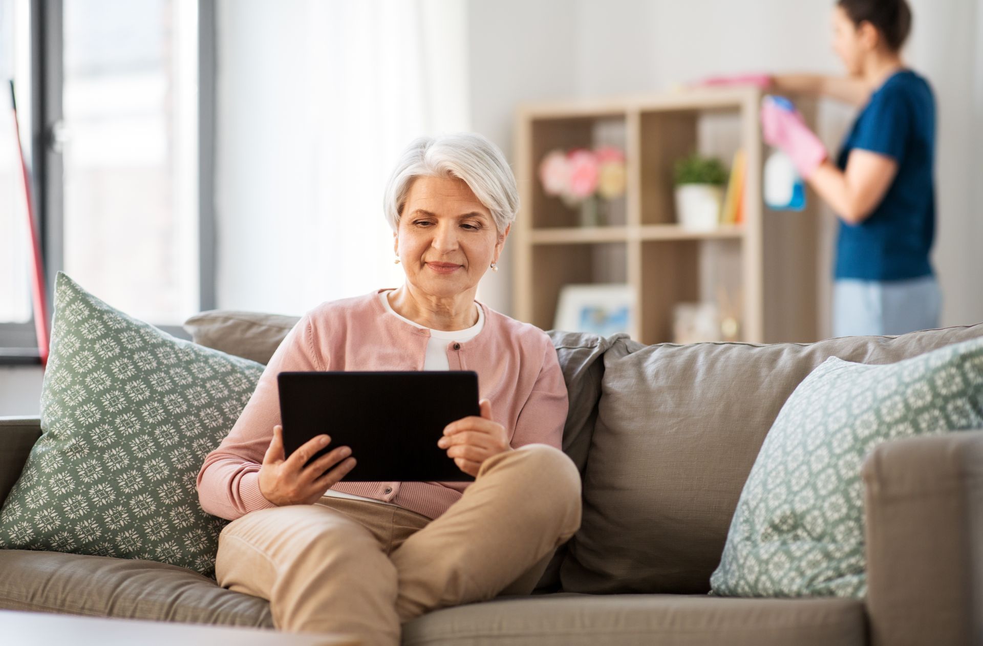 Femme âgée avec une tablette et personnel de ménage en arrière-plan.