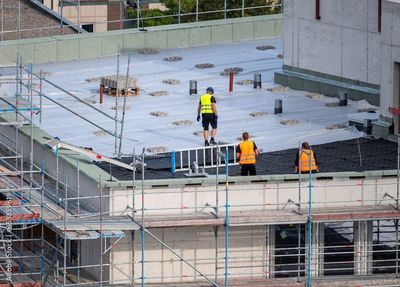Bauarbeiter auf einem Gebäudedach. Einer in gelber Weste geht, zwei in orangefarbenen Westen arbeiten. Um das Gebäude herum ist ein Gerüst.