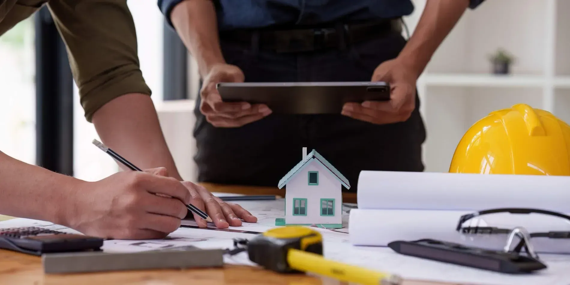Dos personas revisan planos y una maqueta de casa en un escritorio con herramientas de construcción y un casco amarillo cerca.