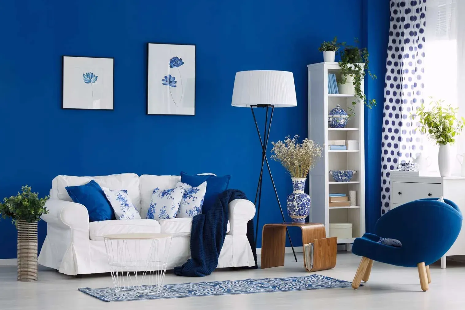 Un sofá blanco con cojines azules descansa contra una pared azul vibrante en una habitación con una lámpara de pie y una estantería blanca.