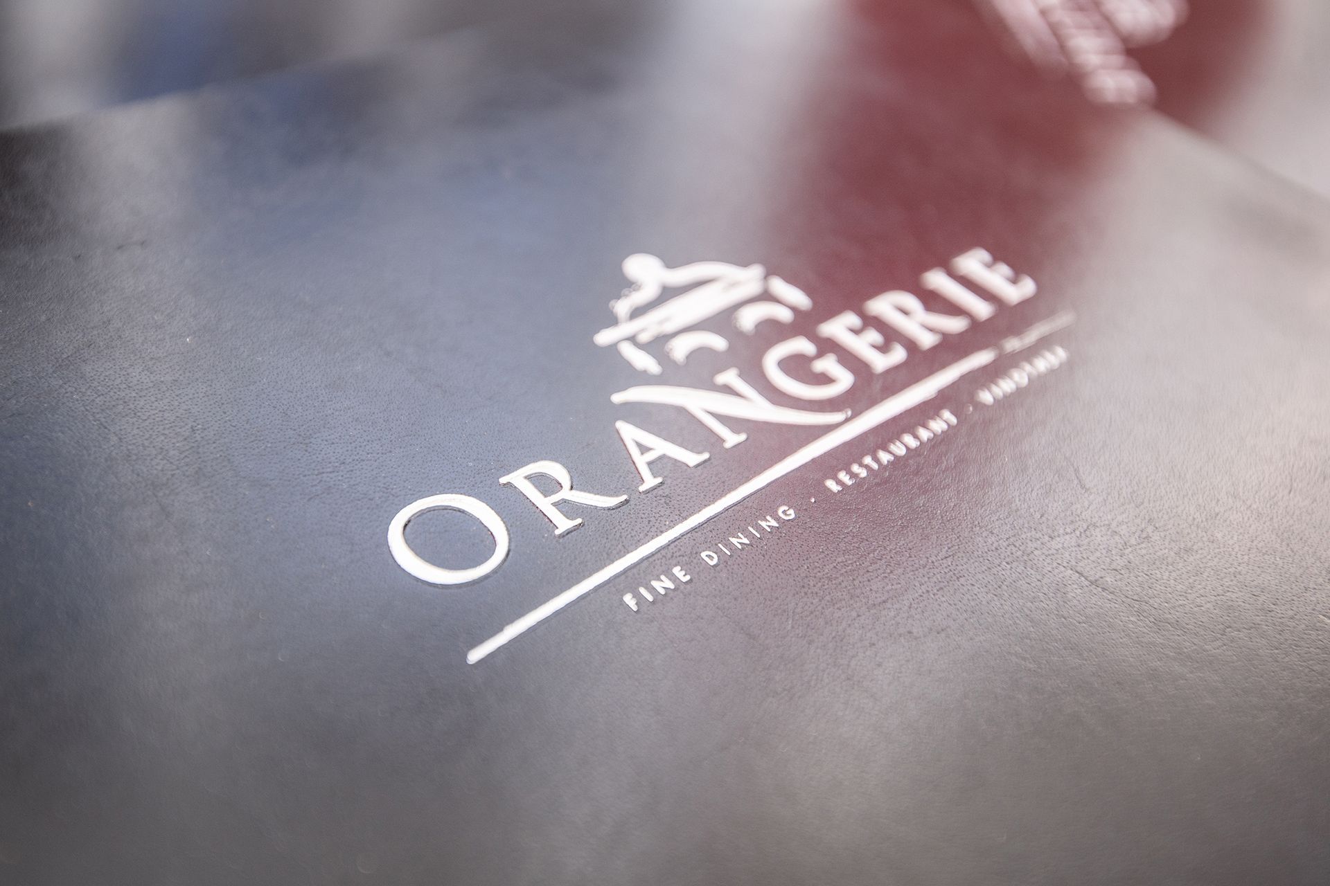 Weißes ORANGERIE-Logo auf dunklem, strukturiertem Untergrund, schräg betrachtet.